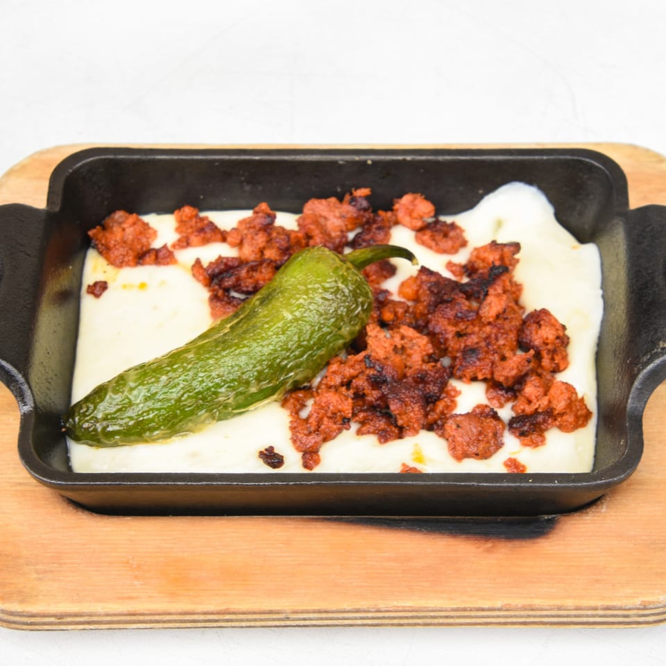 Melted white cheese with chorizo and a green pepper in a black skillet on a wooden board.
