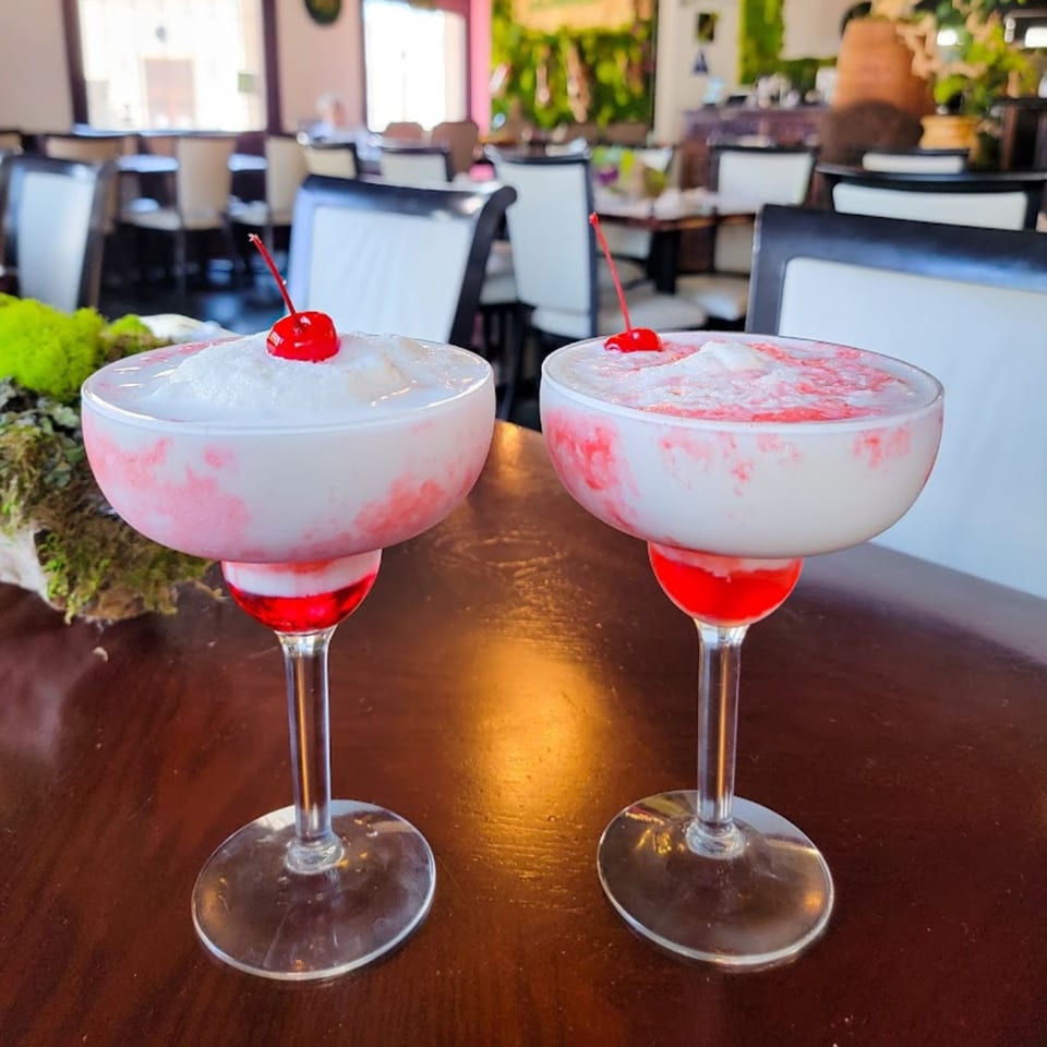 Two frozen margaritas with white tops and red swirls, garnished with cherries, on a wooden table.