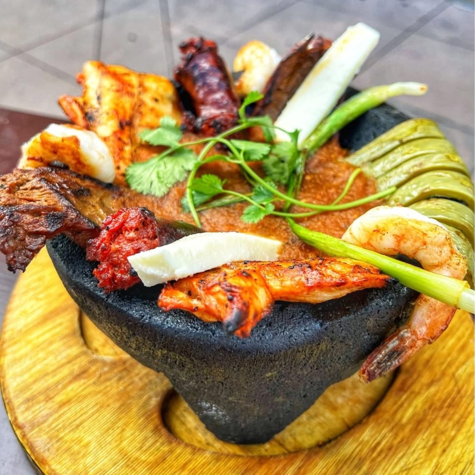 A molcajete filled with grilled meats, shrimp, cheese, nopales, and cilantro on a wooden base.