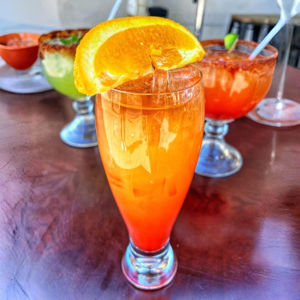 A vibrant orange and red cocktail with an orange slice, alongside other Mexican drinks on a table.