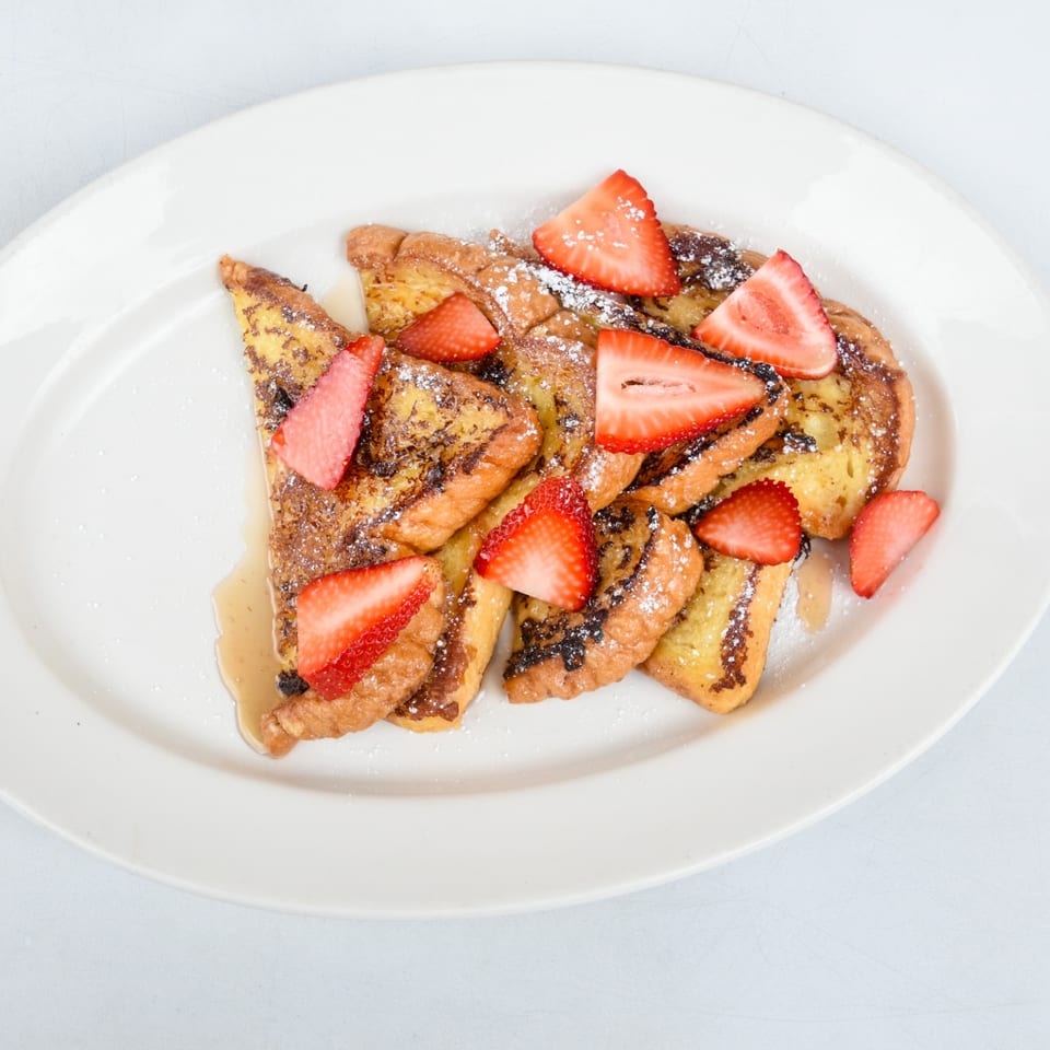 Golden French toast triangles with powdered sugar, fresh strawberries, and maple syrup on a white plate.