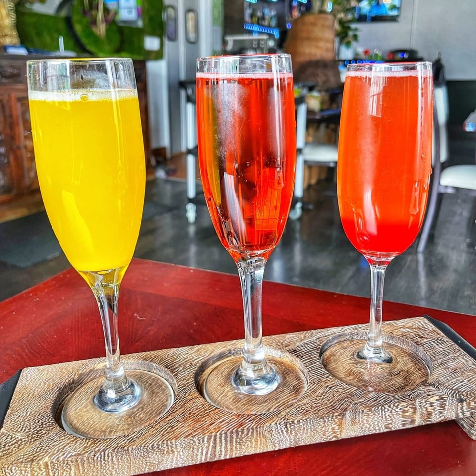 Three colorful mimosa drinks, yellow, red, and orange, on a wooden tray at Lunas Bar and Grill.