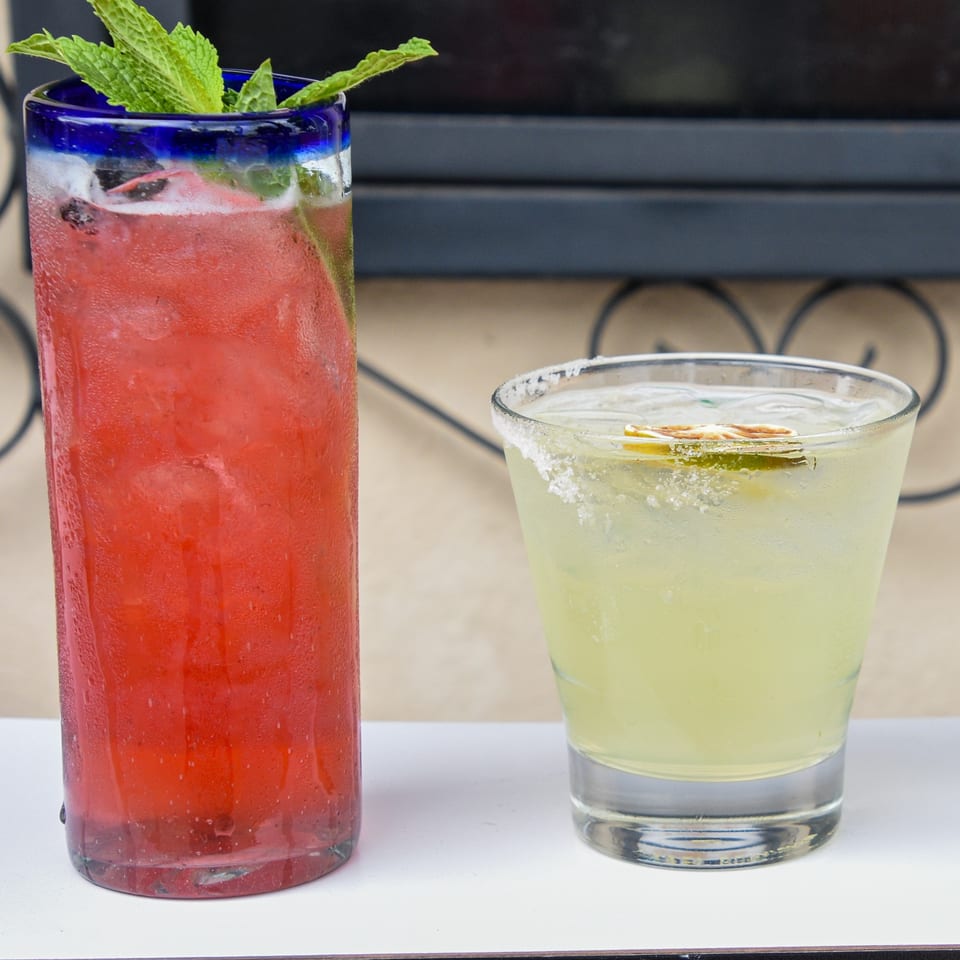 Two refreshing drinks, a tall red cocktail with mint and a lime margarita, on a white surface.