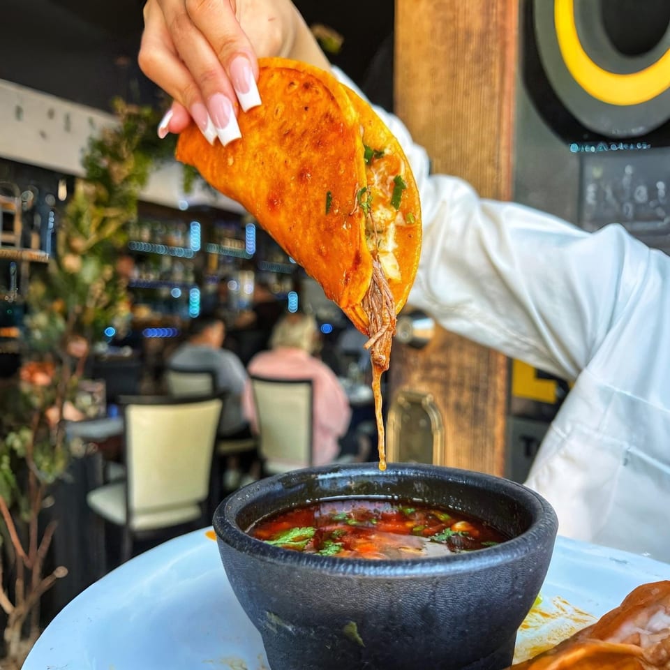 Hand holding a birria taco dripping consommé into a black bowl at Lunas Bar and Grill.