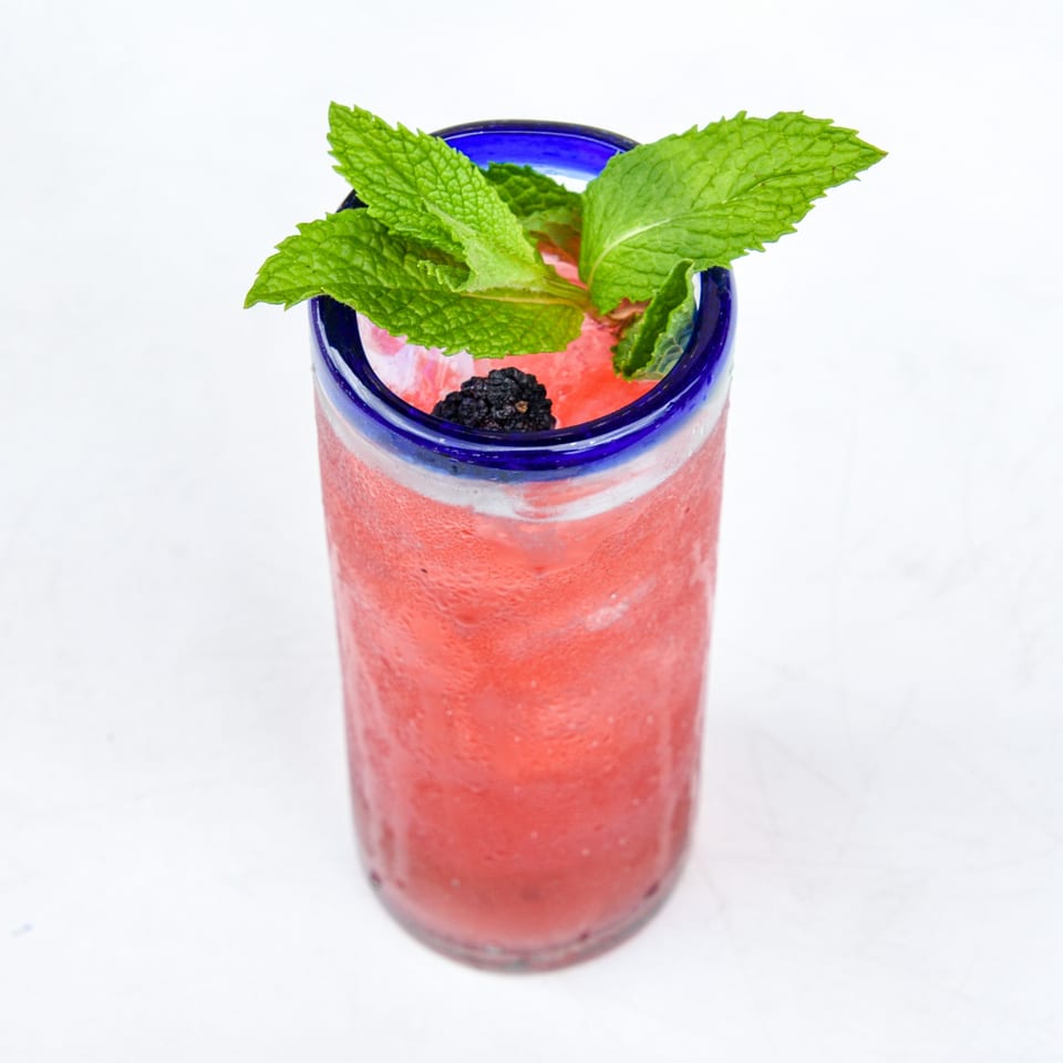 A vibrant red frozen drink in a blue-rimmed glass, garnished with fresh mint and a blackberry.