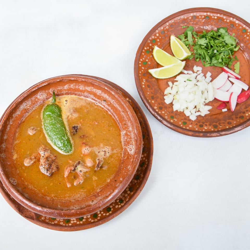 Mexican soup in a clay bowl with a green chili, served with a side of limes, cilantro, onions, and radishes.