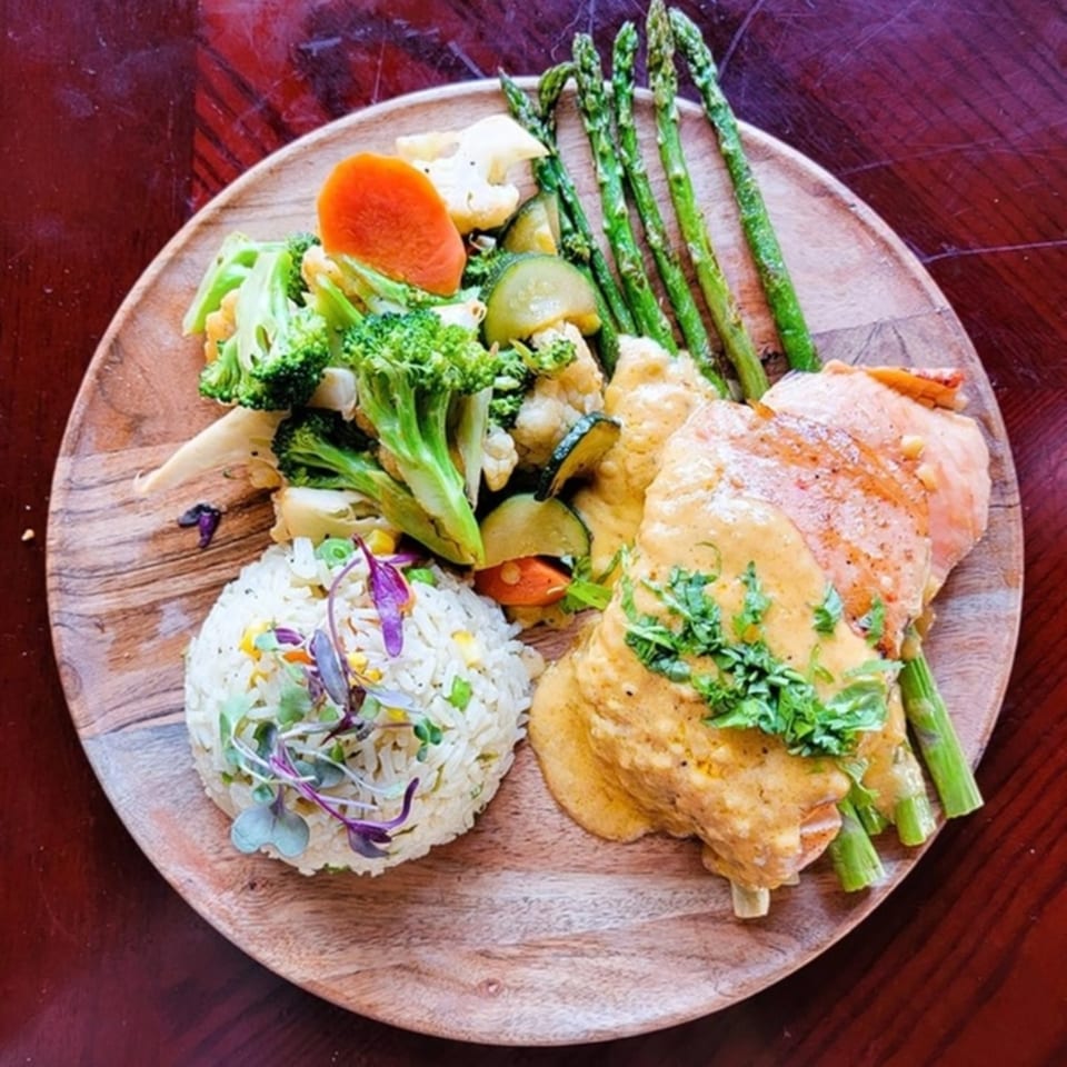 Salmon with yellow sauce, rice, asparagus, broccoli, and other vegetables on a wooden plate.